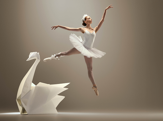 Ballerina performing a jump next to a large origami swan structure, symbolizing physical lightness and freedom of movement